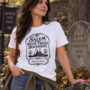 May include: A white short-sleeved t-shirt featuring a black graphic print that reads Salem Witch Trials Descendant 1692 Rebecca Nurse, accompanied by an illustration of a house, trees, and birds within an ornate decorative frame.