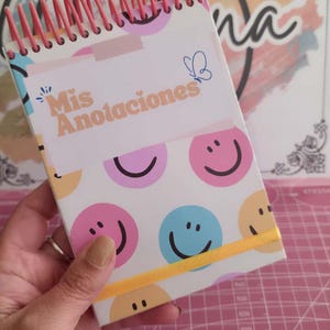 Puede incluir: Un bloc de notas con espiral rosa y banda elástica amarilla. La portada presenta el texto "Mis Anotaciones" y caras sonrientes en rosa, azul y amarillo. También hay un gráfico de mariposa azul.