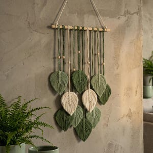 Puede incluir: Un tapiz de pared de macramé con diseños de hojas verdes y blanquecinas. Las hojas están suspendidas de una varilla de madera con hilo de color natural y cuentas de madera. La decoración de la pared está colgada de una cuerda de color neutro.