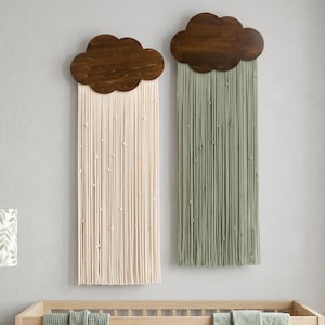 May include: Two cloud-shaped wall hangings with wooden tops and hanging strands. One is cream-colored, the other is sage green. The hangings are above a wooden crib with a sage green blanket. A lamp with a leaf pattern is on the left.