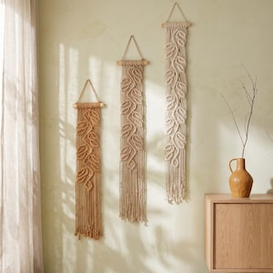 May include: Three vertical macrame wall hangings suspended from wooden dowels with triangular hanging cords. Each piece features a central knotted leaf pattern with long, loose fringe at the bottom. The colors range from tan to cream.