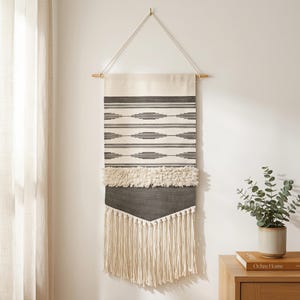 May include: A rectangular wall hanging featuring a cream and charcoal gray woven pattern with horizontal stripes and diamond motifs. The piece includes a textured shaggy section, a solid dark gray panel, and long cream fringe at the bottom.