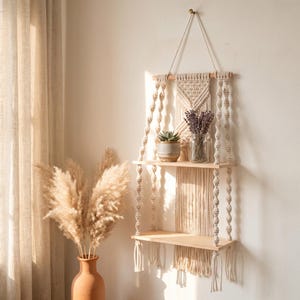May include: A two-tiered wall-mounted shelf featuring light-colored wooden planks suspended by intricate off-white macrame cord with knotted patterns. The structure hangs from a single point and includes a small potted succulent and a glass jar.
