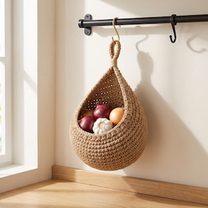 May include: A teardrop-shaped hanging basket made of thick, tan-colored woven jute cord. The basket is suspended from a gold-toned metal hook on a black horizontal bar and contains two red onions, one yellow onion, and a head of white garlic.