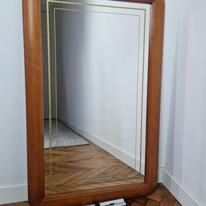 May include: A rectangular wall mirror with a thick, rounded-edge medium brown wooden frame. The glass surface features a thin, double-lined gold rectangular border etched into the center. The mirror is positioned upright on a wooden floor.