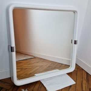 May include: A square white framed mirror with rounded corners. The mirror has two silver metal brackets on the sides. The mirror is leaning against a white wall and a wooden floor.