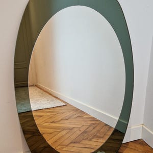 May include: An oval-shaped mirror with a wide, dark olive green tinted glass border surrounding a clear central reflective surface. The mirror is resting on the floor, partially overlapping a white cloth with the printed text LIFE visible.