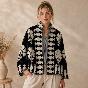 Könnte beinhalten: Eine schwarze Samtjacke mit aufwendigen cremefarbenen Blumen- und Geometriestickereien auf den Vorderteilen und Ärmeln, getragen über einem schwarz-weiß gestreiften Hemd und einer hellen Hose.