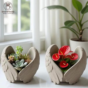 May include: Two beige angel wing-shaped planters, one filled with succulents and the other with red anthurium flowers. The planters are on a white surface near a window. The text "DIGITBYASAD CRAFT & SUPPLIES" is visible.
