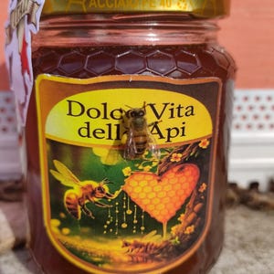 May include: A jar of honey with a gold lid and a label featuring a bee illustration and the words "Dolce Vita dell'Api." A bee is perched on the jar. The honey is a dark amber color.