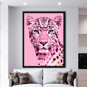 以下が含まれることがあります： ピンク色のヒョウの肖像画が描かれた額入りのアートプリント。ヒョウは黒い斑点と緑色の目を持ち、ピンク色の背景に配置されています。このアートワークは、装飾的なクッションが付いた灰色のソファの上に飾られています。