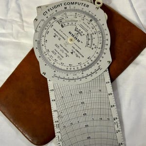May include: A vintage C1 Flight Computer with a circular slide rule and a rectangular grid, resting on a brown leather case. The computer features scales for time, speed, distance, and fuel computation. The text "RESEJUTE" is visible.