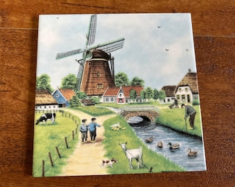 May include: A square ceramic tile featuring a colorful illustration of a rural landscape with a large windmill, a stone bridge over a stream, a grazing cow, a goat, ducks in the water, and two children walking along a dirt path.