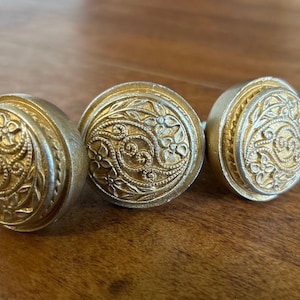 May include: Three circular metal knobs featuring a raised gold-toned floral and scrollwork pattern on the face. Each knob has a smooth, matte silver-toned outer rim and a textured, intricate center design with small flowers and swirling vine details.