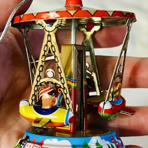May include: A miniature metal carousel ornament featuring colorful hanging airplane seats with painted figures. The structure has a red and gold roof, a central pillar, and a base decorated with a landscape scene of mountains, trees, and buildings.