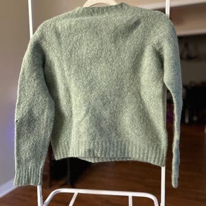 May include: A sage green long-sleeve sweater with a crew neckline and a textured, fuzzy knit finish. The garment features ribbed cuffs at the wrists and a ribbed hemline at the bottom, providing a structured fit for this solid-colored pullover.