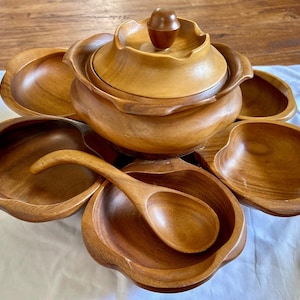 May include: A wooden serving set consisting of a large central lidded bowl with a knob handle, surrounded by four smaller petal-shaped bowls. The set includes a matching wooden ladle resting inside one of the bowls, all featuring a smooth, polished finish.