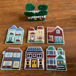 May include: A collection of seven plastic canvas needlepoint pieces featuring six flat house designs in various colors and one three-dimensional garden fence structure with green trees, all crafted with detailed cross-stitch patterns.