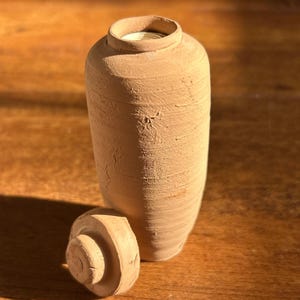 May include: A small, cylindrical, light brown terracotta jar with a textured, matte surface and a matching circular lid resting beside it. The jar features a narrow opening at the top and a simple, unglazed finish with visible clay striations.