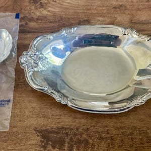 May include: A silver serving dish with ornate edges and a matching spoon in a plastic sleeve. The spoon has a decorative handle. The dish and spoon are on a wooden surface.
