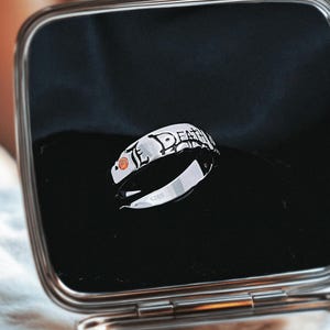 May include: A silver ring with the word "DEATH" in black lettering. The ring has a small orange gemstone and is displayed in a black box.
