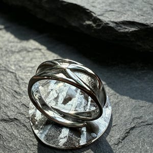 May include: A silver-toned ring featuring a polished, crisscross band design with a textured, matte finish on the inner sections. The metal band is open at the back, and the interior surface clearly displays the engraved hallmark S925.