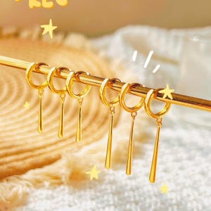 Könnte beinhalten: Goldfarbene Creolen mit hängenden, länglichen Tropfenanhängern. Die Ohrringe werden auf einem goldenen Stab vor einem weichen, strukturierten Hintergrund präsentiert. Die Ohrringe sind hellgoldfarben.