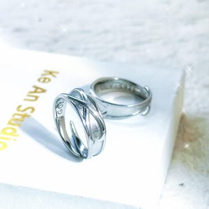 May include: Two silver rings. One ring features an interwoven design, while the other is a simple band. The rings are displayed on a white surface with the text "Ke An Studio" visible in gold.