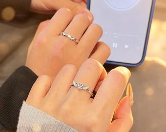 May include: Two silver-toned rings worn on fingers. One ring features a central heart-shaped stone flanked by small wings. The other ring is a simple band with a small embedded stone. The rings are metallic with a polished finish and clear stones.