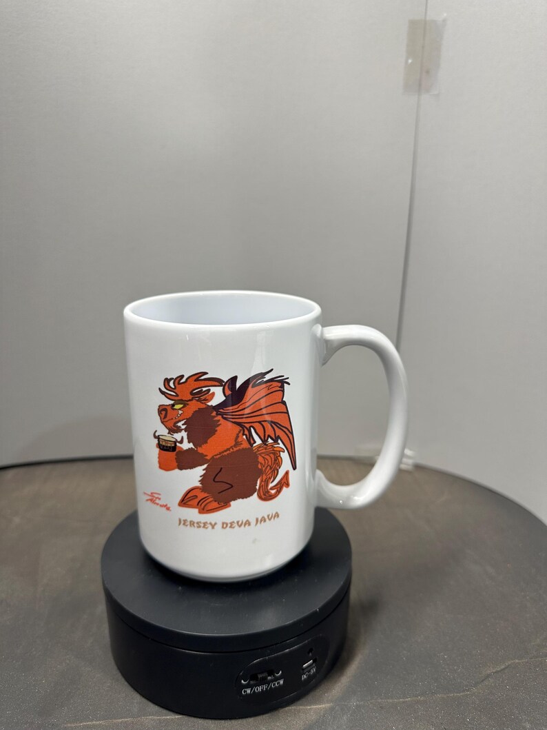 May include: White ceramic mug with a cartoon winged creature in brown and orange, holding a coffee cup. The mug has the text "Jersey Deva Java" and a signature. The mug is on a black rotating stand.