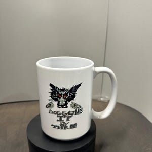 May include: White ceramic mug with a black and red graphic of a creature and the text "Dog Gone It By The Tree". The mug has a curved handle and is on a black rotating stand.