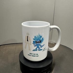 May include: White ceramic mug with a cartoon design. It features two figures and the text "DON'T FIT IN, STICK RIGHT OUT." One figure is a stick figure in a suit, the other is a blue character with spiky hair. The mug has a curved handle.