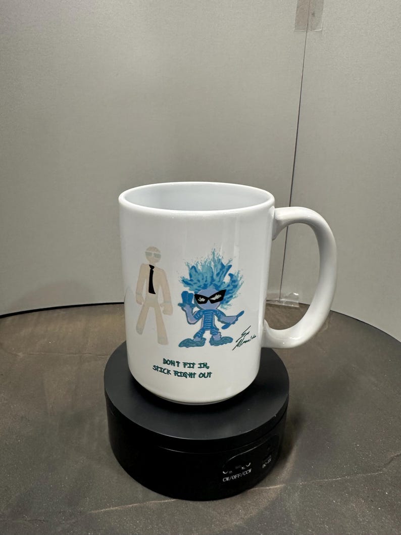 May include: White ceramic mug with a cartoon design. It features two figures and the text "DON'T FIT IN, STICK RIGHT OUT." One figure is a stick figure in a suit, the other is a blue character with spiky hair. The mug has a curved handle.