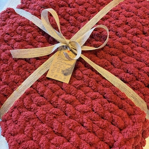 May include: A deep red chunky knit blanket made of thick yarn, folded and tied with a light tan ribbon. A small rectangular cardboard tag is attached to the ribbon, featuring care instructions and the text 100% Polyester.