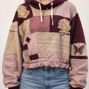 Pode incluir: Um moletom patchwork em tons de bordô, bege e rosa. O moletom cropped apresenta aplicações florais e de borboletas, capuz com cordão e cintura franzida. As mangas compridas têm punhos bordô. A modelo está usando jeans azuis.
