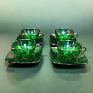 Può includere: Un set di quattro tazze da tè in vetro verde smeraldo con piattini quadrati abbinati. Ogni tazza presenta una forma quadrata arrotondata con un piccolo manico, e i piattini hanno un design quadrato con un incavo centrale per la base.