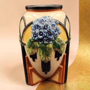 May include: A ceramic vase with a blue hydrangea flower and green leaves. The vase has a cream-colored background with black and orange geometric designs. The top rim is black.