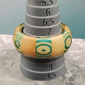Puede incluir: Un brazalete de madera con un diseño marrón claro y verde azulado. El brazalete se coloca en una herramienta de medición gris. El brazalete tiene un diámetro de aproximadamente 7 cm.