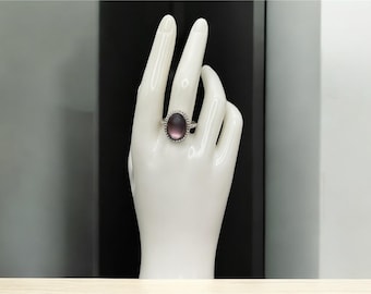 Puede incluir: Un anillo de plata con una gran piedra preciosa ovalada de color púrpura. El anillo se muestra en una mano de maniquí blanca. La piedra preciosa está rodeada por un borde de cuentas. El anillo es una pieza de joyería de moda. El fondo es una mezcla de blanco y negro.