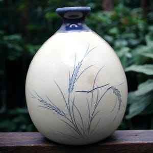 Puede incluir: Jarrón de cerámica color crema con borde azul y cuerpo redondeado. El jarrón presenta una delicada ilustración azul de espigas de trigo. El fondo está borroso, lo que sugiere un entorno al aire libre.