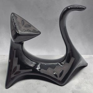 May include: A glossy black ceramic cat figurine with a stylized, abstract form. The sculpture features a pointed head, an arched tail, and matte gray geometric patterns on the ears, body, and base, creating a sharp, angular silhouette.