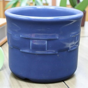 Puede incluir: Una maceta de cerámica azul con un diseño texturizado, similar a un tejido, alrededor del centro. La maceta tiene una forma redondeada y un acabado liso y brillante. Ideal para plantas pequeñas o suculentas.