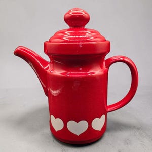 Può includere: Una teiera in ceramica rosso brillante con finitura lucida, coperchio abbinato con pomello arrotondato, beccuccio curvo e manico laterale. La base della teiera è decorata con un motivo ripetuto di cuori bianchi che circonda la parte inferiore.