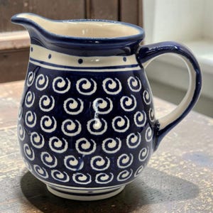 Puede incluir: Una jarrita de cerámica con esmalte azul profundo y un patrón repetitivo de espirales blancas pintadas a mano. La jarra cuenta con un asa curva, un pico vertedor y un acabado liso y brillante con interior y base de color blanco.