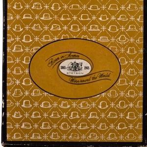 May include: A rectangular box with a mustard yellow background featuring a repeating white diamond pattern containing small hat icons. A central oval label displays the text Born in America, 1865-1965, Stetson, and Worn round the World.
