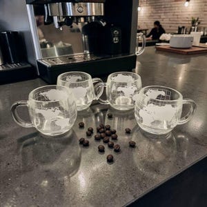 May include: Four clear glass coffee mugs with a world map design are arranged on a countertop. Dark coffee beans are scattered in front of the mugs. An espresso machine is in the background.