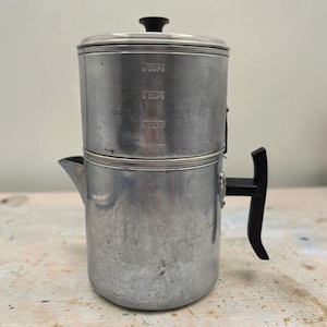 Puede incluir: Una cafetera de metal plateado con asa y pomo de tapa negros. La sección superior presenta marcas de medición en relieve para 2, 4, 6 y 8 tazas. El cuerpo cilíndrico tiene un pico vertedor y un asa negra vertical y resistente.
