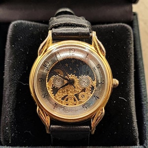 May include: A gold-toned Fossil watch with a black leather band. The watch face has a transparent center revealing the inner workings and a dark blue background with stars and a crescent moon. The watch is in a black box.