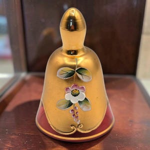 以下が含まれることがあります： 滑らかで光沢のある金色のベル。ベルの前面には手描きの花のデザインが施されており、ピンク色の縁取りがある白い花、緑の葉、小さな紫色のつぼみがベルの中央に描かれています。