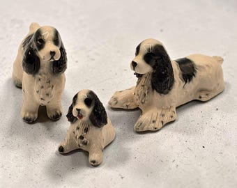 Pode incluir: Três figuras de cerâmica de cães spaniel com uma base de cor creme e manchas pretas nas orelhas e no corpo. Uma figura está de pé, uma sentada no centro e outra deitada. Cada cão tem traços faciais detalhados e um acabamento brilhante.